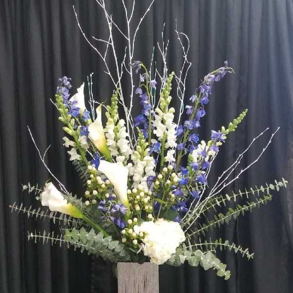 Tall floral arrangement with white calla lilies and blue-purple spikes in a wooden vase