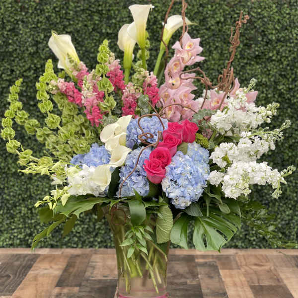 Tall mixed bouquet with pink roses, blue hydrangeas, white calla lilies, and orchids in a glass vase