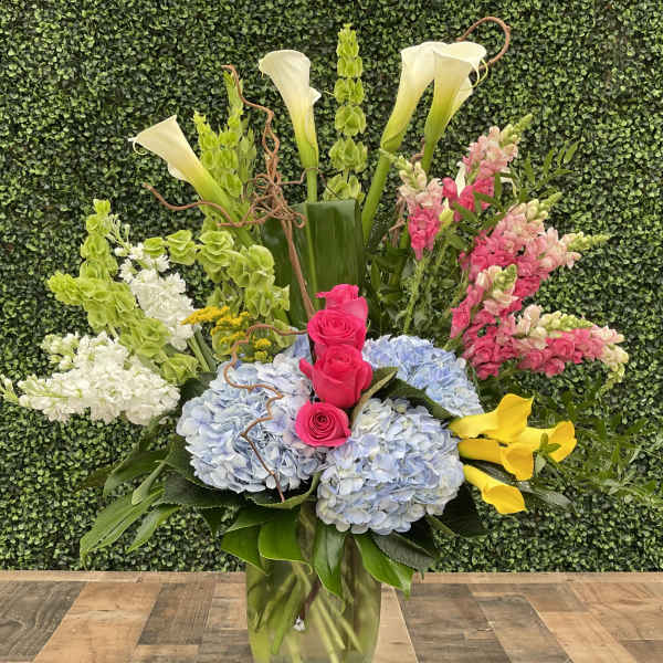 Tall mixed floral arrangement in a glass vase with hydrangeas, roses, calla lilies, and snapdragons