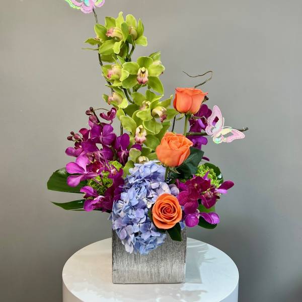 Tall arrangement of green and purple orchids, orange roses, and blue hydrangea in a square vase with butterfly picks