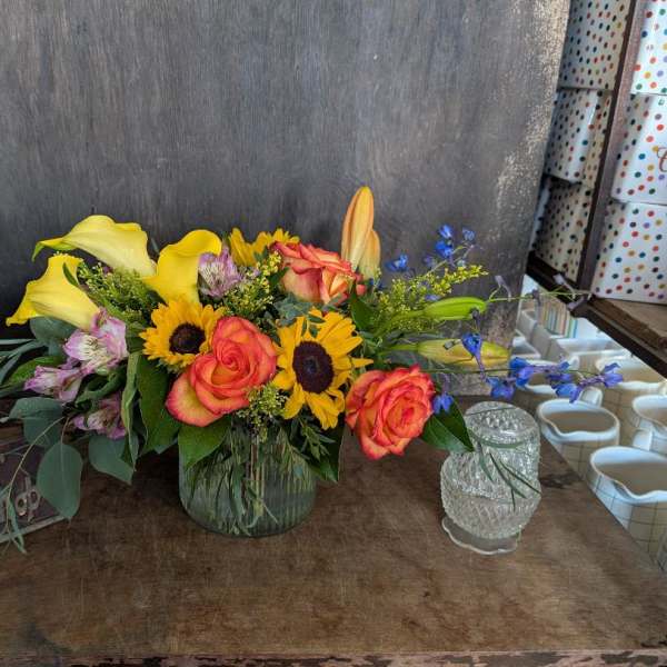 Mixed bouquet with roses, sunflowers, calla lilies, and blue accents in a glass vase