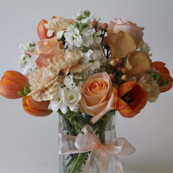 Peach and white bouquet with roses, tulips, and calla lilies in a glass vase with ribbon