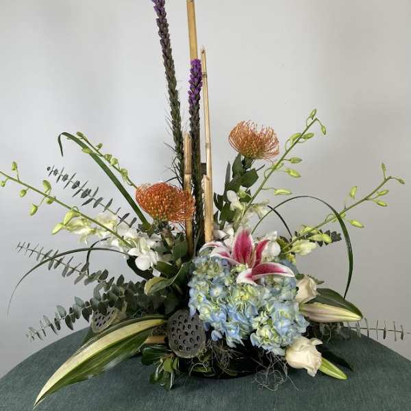 Tall floral arrangement with blue hydrangeas, lilies, and orange pincushion blooms
