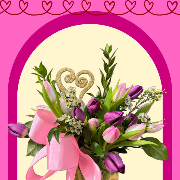 Pink and purple tulip bouquet in a glass vase with a ribbon and heart topper