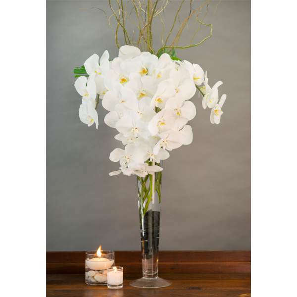 White orchids in a tall glass vase beside two lit candles.