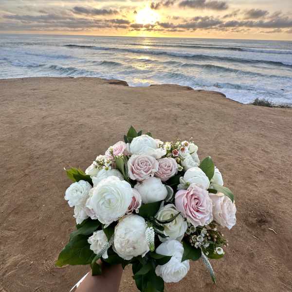 Bridal bouquet of white and blush roses and ranunculus held by the ocean at sunset