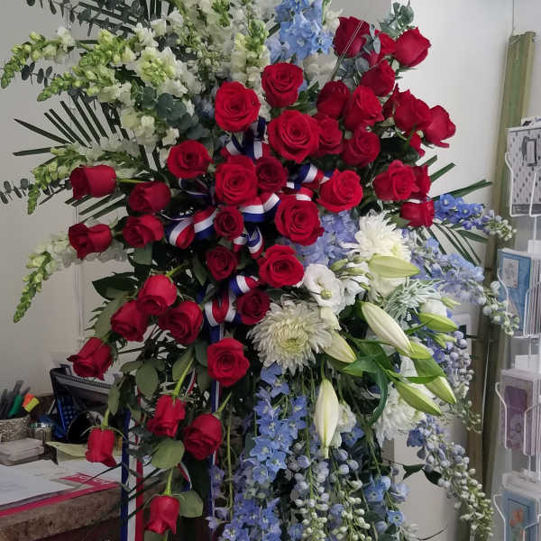 Tall standing spray with red roses, white lilies and mums, blue delphinium, and patriotic ribbon accents