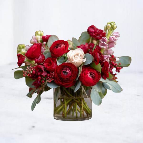 Low glass vase of red ranunculus and roses with a single pale bloom and pink accents.
