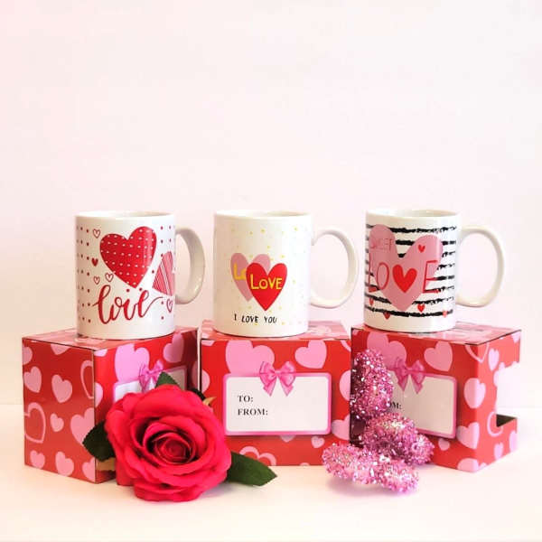 Three Valentine mugs on heart-patterned gift boxes with a red rose and glitter heart.