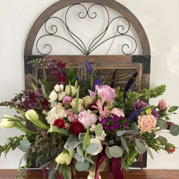 Large mixed flower arrangement in a gold pedestal vase with burgundy ribbon