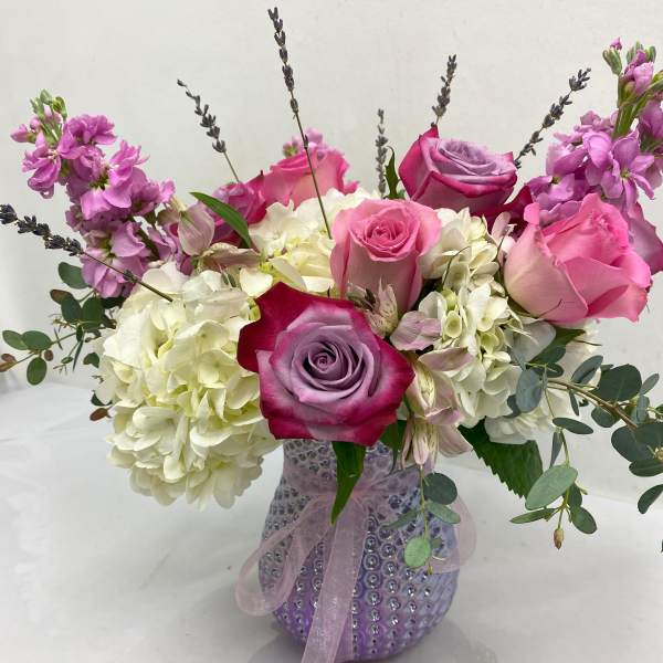 Pink and lavender roses with white hydrangeas in a textured lavender glass vase tied with ribbon