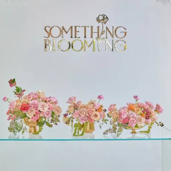 Three pink floral arrangements in gold vases beneath a "Something Blooming" sign