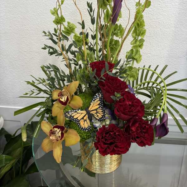Tall floral arrangement with red roses, purple calla lilies, and yellow orchids in a gold vase