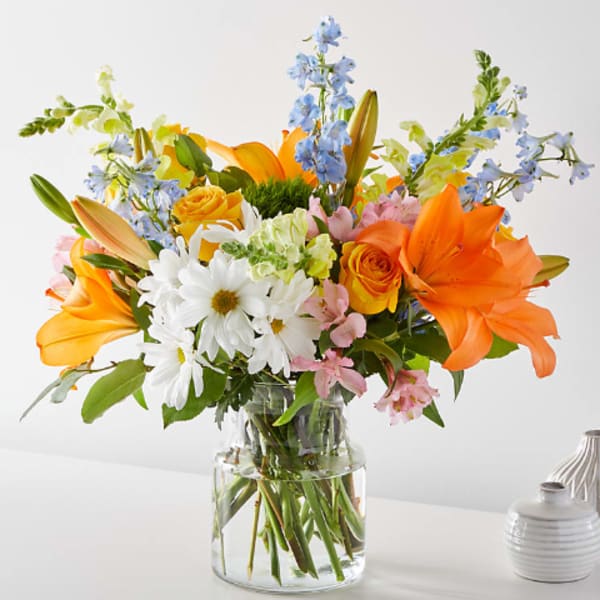 Bright mixed bouquet with orange lilies, yellow roses, and white daisies in a glass vase