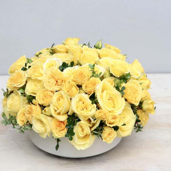 Low round arrangement of yellow roses in a white bowl-shaped container