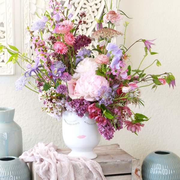 Tall pastel pink and purple flower arrangement in a white ceramic face vase on a rustic crate