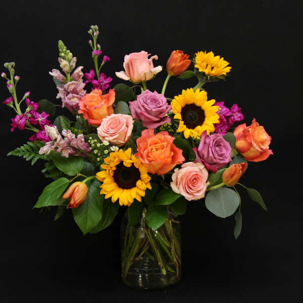 Mixed bouquet of roses, sunflowers, and purple blooms in a glass vase
