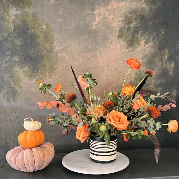Orange floral arrangement in a striped vase beside stacked decorative pumpkins
