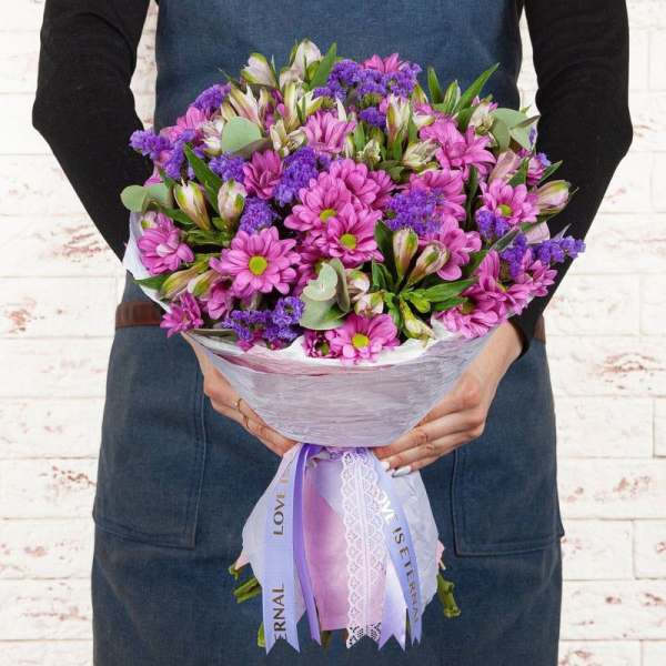 Handheld bouquet of pink daisies and purple filler flowers wrapped in lavender paper