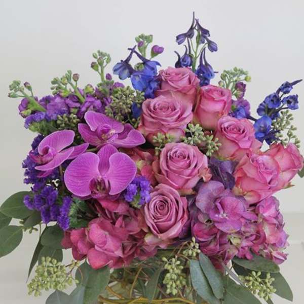 Compact arrangement of pink roses, orchids, and hydrangeas with purple and blue accents in a clear glass cube vase