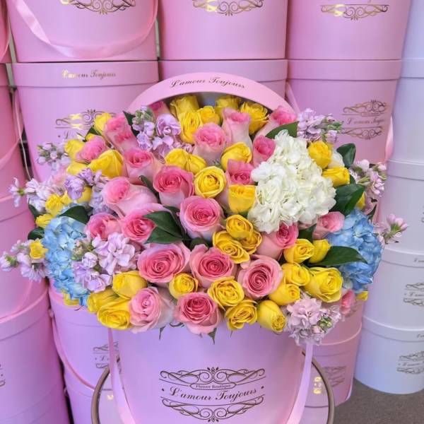 Pink and yellow roses arranged in a large pink hat box with white hydrangeas.