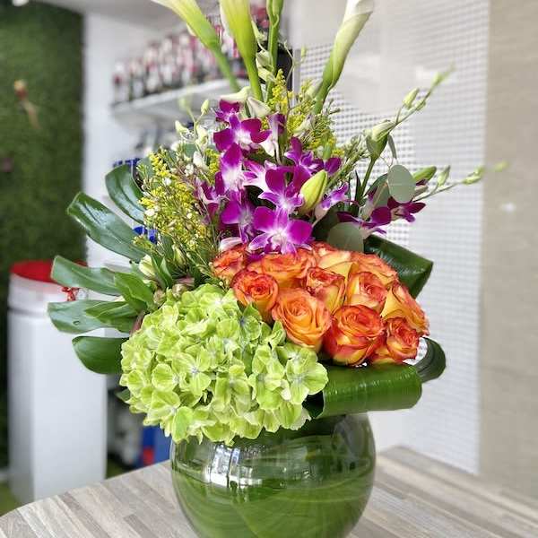 Tall mixed arrangement with calla lilies, orchids, roses, and hydrangea in a leaf-lined glass vase.