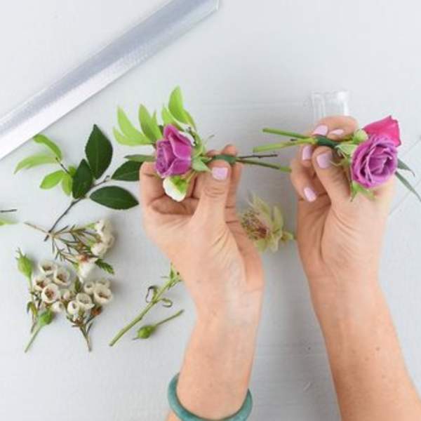 Hands arranging small purple roses with greenery on a white surface