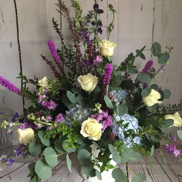 Arrangement of cream roses, blue hydrangeas, and purple spikes in a white vase