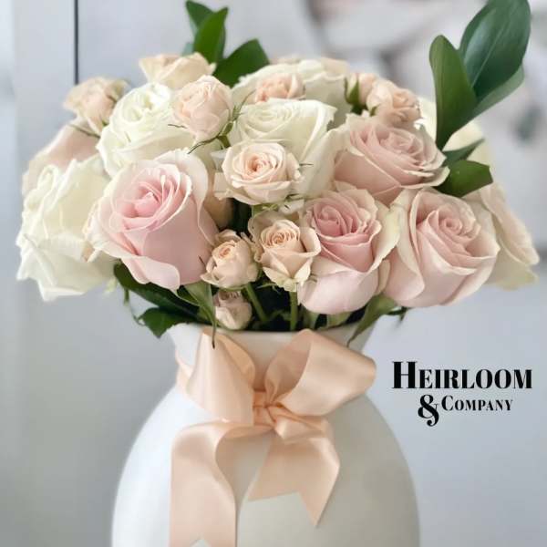 Pink and white roses arranged in a white vase with a peach ribbon