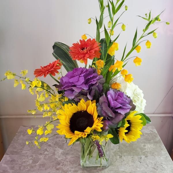 Mixed bouquet with sunflowers, purple kale, and yellow orchids in a glass vase