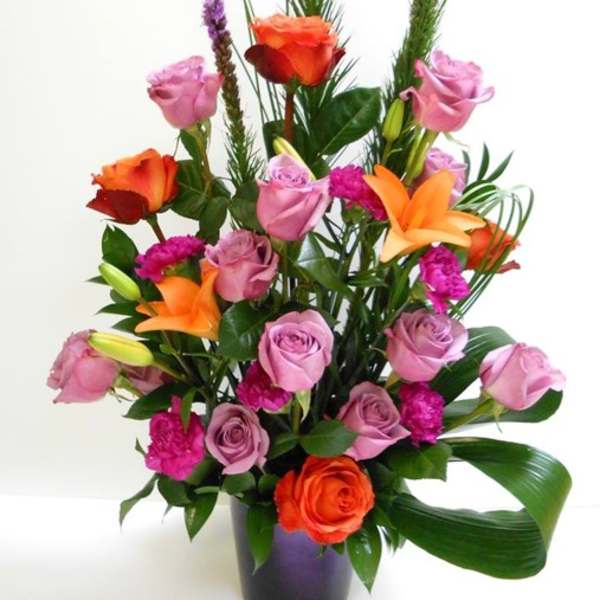 Tall arrangement of lavender roses, orange lilies, and fuchsia blooms in a purple vase with a folded green leaf.