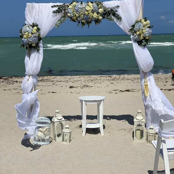 Beach wedding arch with white drapes and blue-and-cream floral arrangements