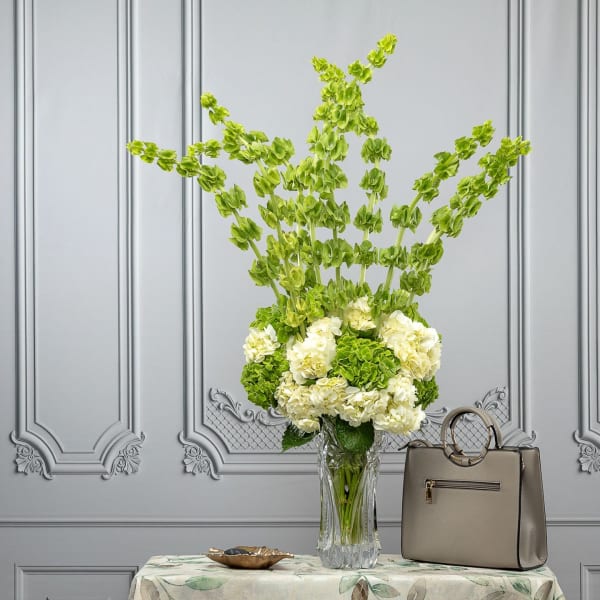 Tall green and white floral arrangement in a clear vase beside a beige handbag