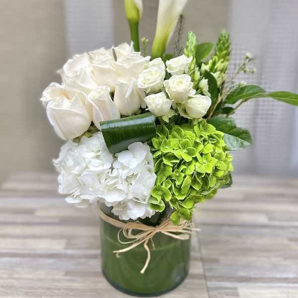 Tall white and green arrangement with calla lilies, roses, and hydrangeas in a glass cylinder vase
