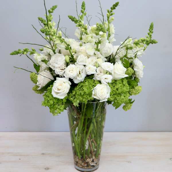 Tall arrangement of white roses and snapdragons with green blooms in a clear glass vase.