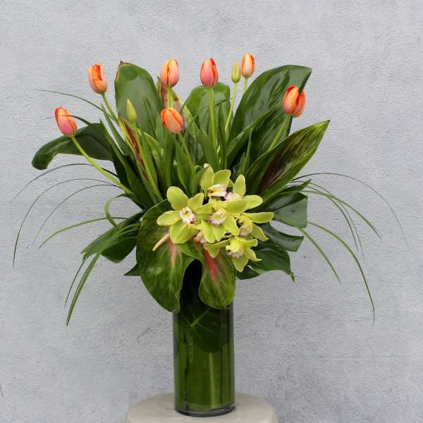 Orange tulips and green orchids in a tall glass vase