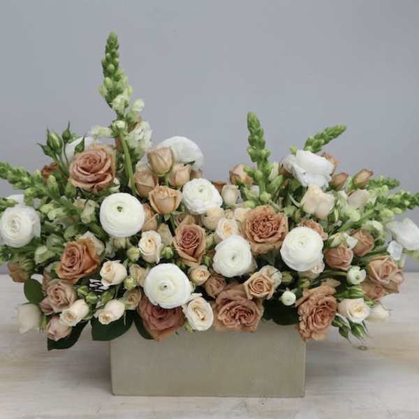 Low rectangular arrangement of white ranunculus and peach roses with snapdragons in a beige container