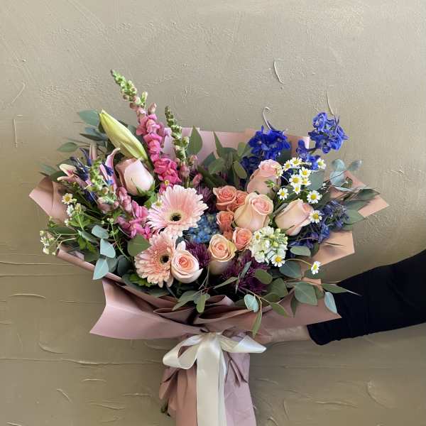 Hand-tied bouquet of pink roses, gerbera daisies, and blue flowers in pink wrap