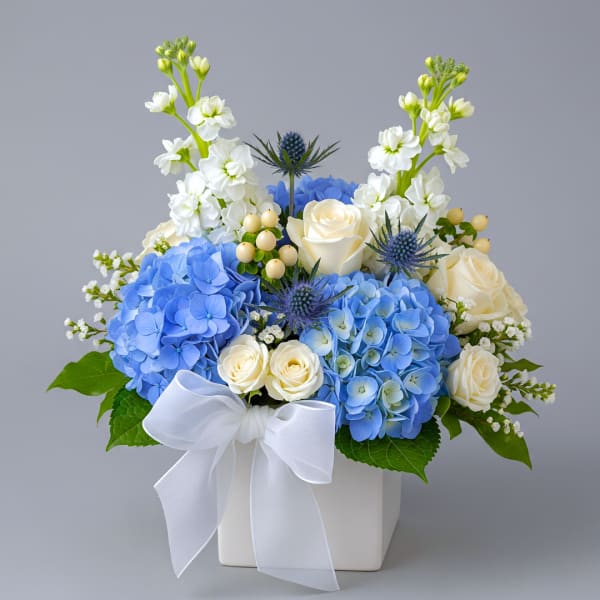 Blue hydrangeas and white roses in a white square vase with a bow