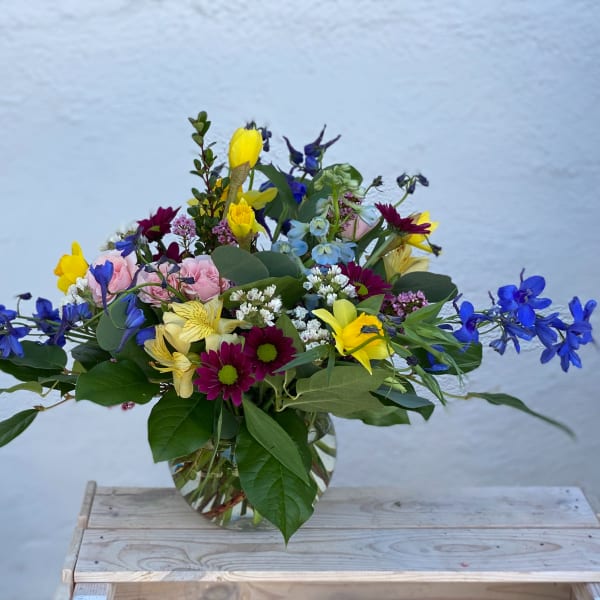 Mixed bouquet in a clear glass vase with blue, yellow, pink, and purple flowers.