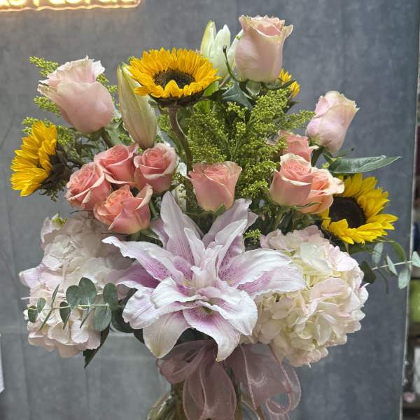 Tall mixed bouquet of pink roses, lilies, hydrangeas and yellow sunflowers in a clear glass vase with ribbon