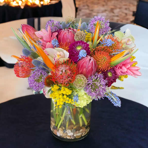Bright mixed tropical and protea flower arrangement in a clear glass vase with stones.