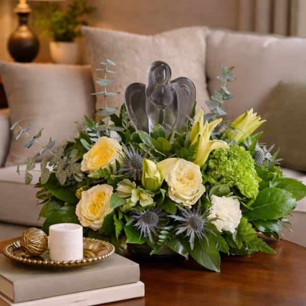 Low arrangement of yellow roses and lilies with a glass angel figurine on a table