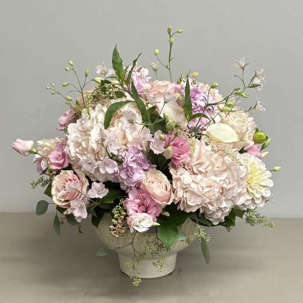 Low arrangement of blush and pink roses, hydrangeas, and mixed blooms in a white pedestal bowl