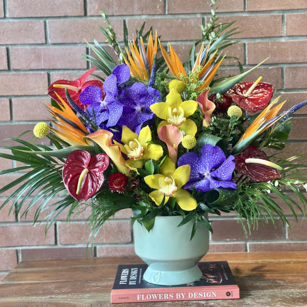 Tall tropical arrangement of orange, purple, yellow, and red flowers in a pale green vase