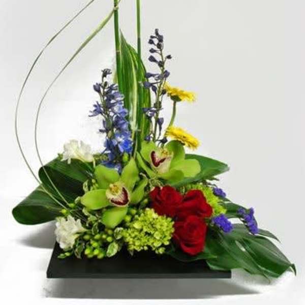 Tropical floral arrangement with red roses, green orchids, and blue flowers on a black base