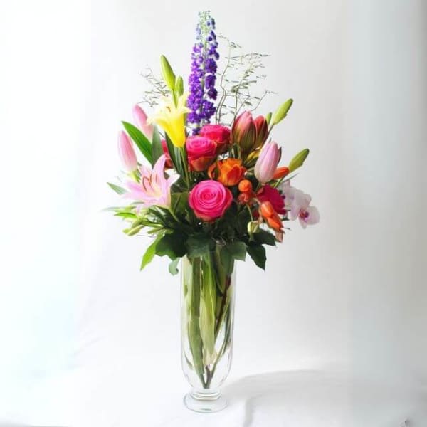 Tall mixed bouquet of pink roses, lilies, orange blooms and purple spikes in a clear glass vase