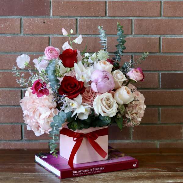 Mixed bouquet of roses, peonies, and hydrangeas in a pink box with a red ribbon