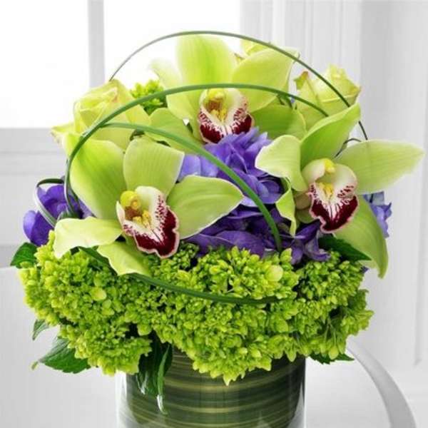 Compact arrangement of green cymbidium orchids and purple flowers in a clear glass cylinder vase