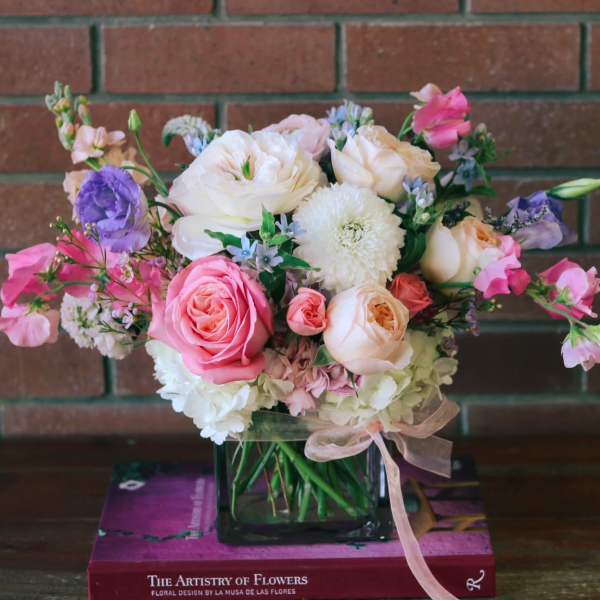 Low pastel arrangement of pink roses and mixed blooms in a square glass vase with ribbon.